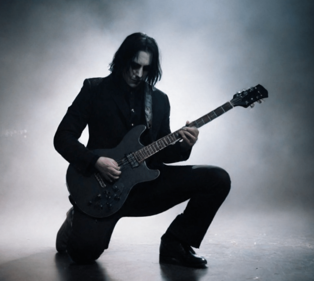 A guitarist in a black suit kneeling on stage, playing a black electric guitar, surrounded by fog and dramatic lighting.